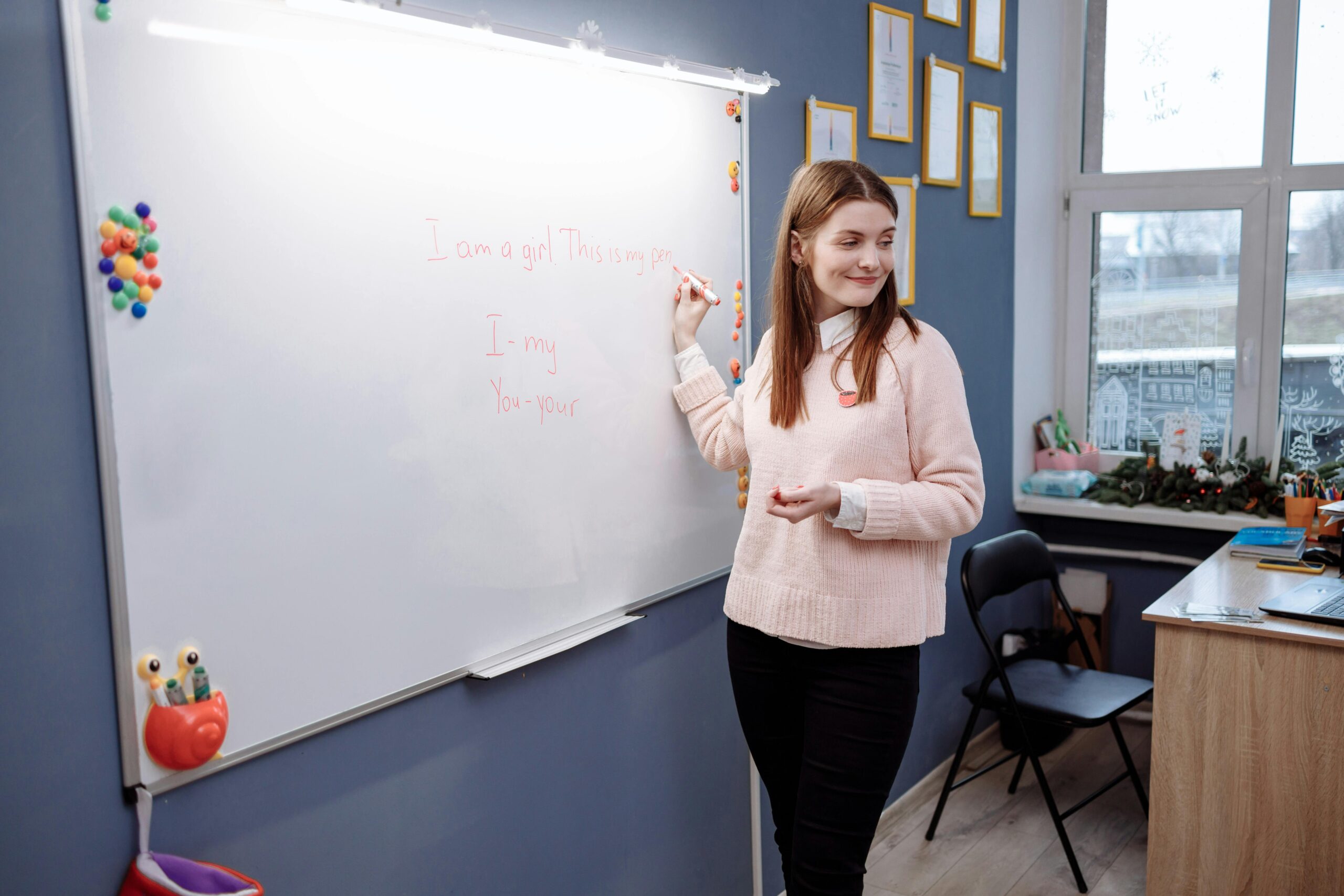 Young woman teaching English lesson in a classroom, writing on a whiteboard.